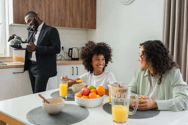 Happy african american mother and daughter looking at each other near businessman talking on smartphone during breakfast — Stock Photo