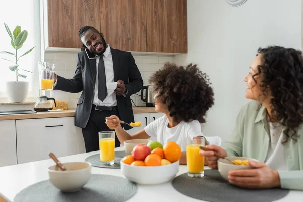 Glücklich afrikanisch-amerikanischer Geschäftsmann hält Krug mit Orangensaft in der Hand, während er auf dem Smartphone spricht und seine Familie betrachtet — Stockfoto