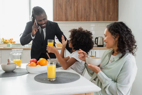 Happy african american girl looking at father in suit talking on smartphone during breakfast — Stock Photo