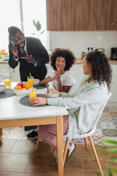 Menina americana africana feliz olhando para a mãe enquanto toma café da manhã perto do pai em terno apontando para o smartphone — Fotografia de Stock