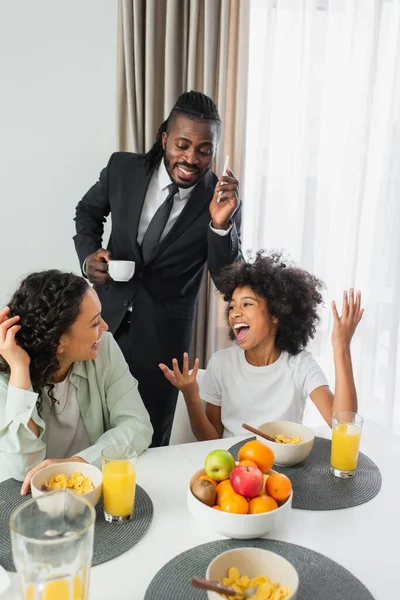 Homem americano africano feliz em terno falando no smartphone e olhando para a família durante o café da manhã — Fotografia de Stock