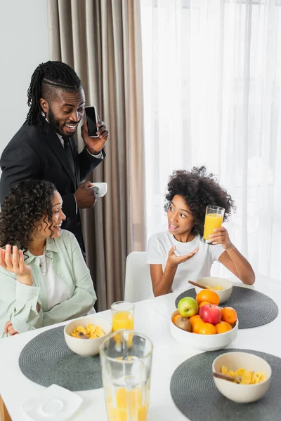 Изумленная африканская американка, указывающая на стакан апельсинового сока рядом с родителями во время завтрака — стоковое фото