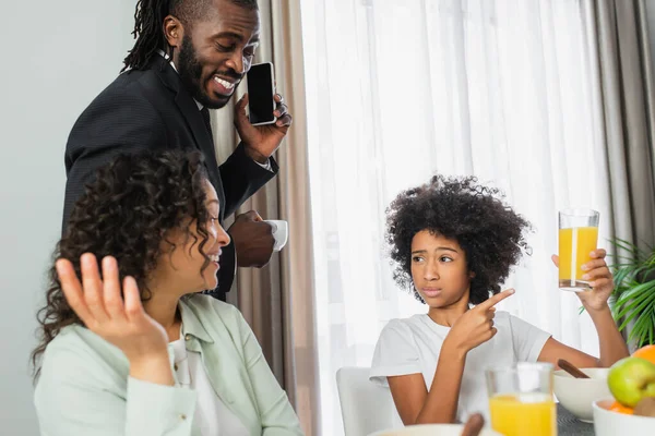Preteen african american girl pointing at glass of orange juice near happy parents during breakfast — Stock Photo