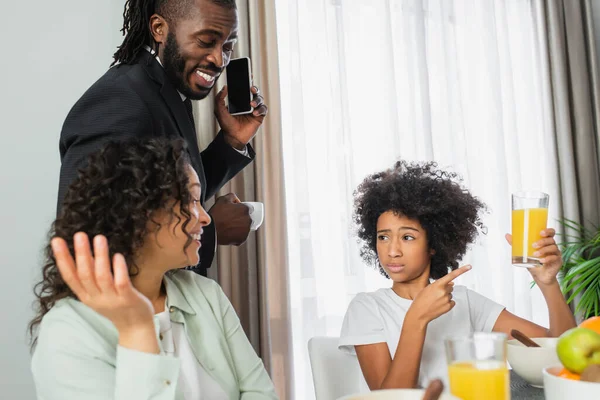 Preteen ragazza afro-americana che indica un bicchiere di succo d'arancia vicino ai genitori felici durante la colazione — Foto stock