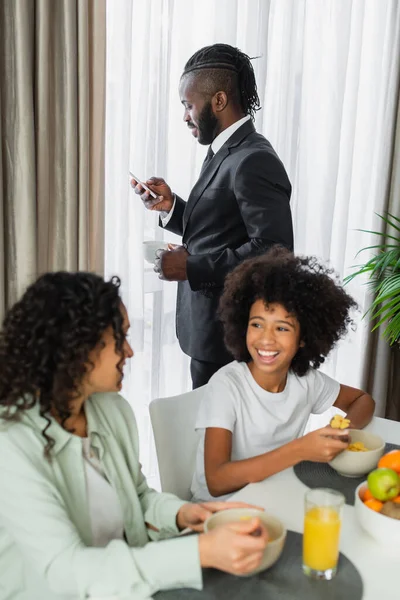 Homem americano africano de terno usando smartphone e segurando xícara de café perto da família tomando café da manhã em primeiro plano desfocado — Fotografia de Stock