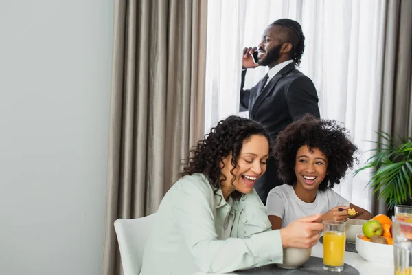 Hombre afroamericano en traje hablando en smartphone cerca de familia feliz desayunando - foto de stock