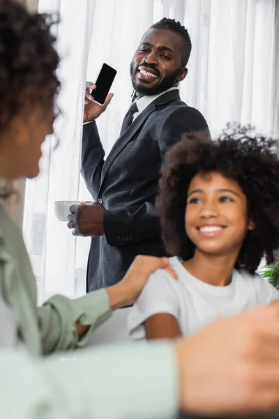 Heureux homme afro-américain en costume tenant smartphone et tasse de café près de la famille sur le premier plan flou — Photo de stock