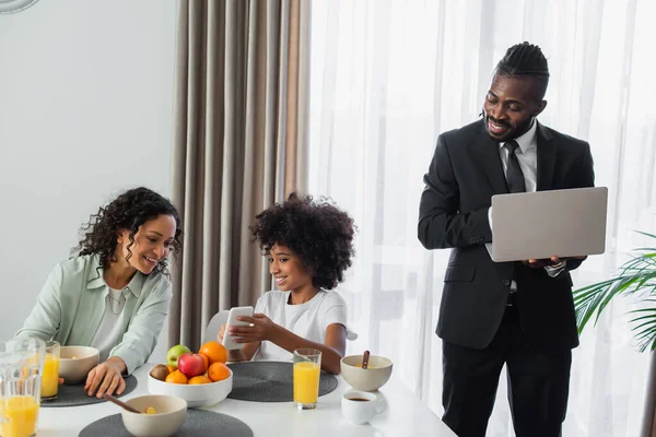 Feliz homem americano africano de terno em pé com laptop enquanto filha mostrando smartphone para a mãe durante o café da manhã — Fotografia de Stock