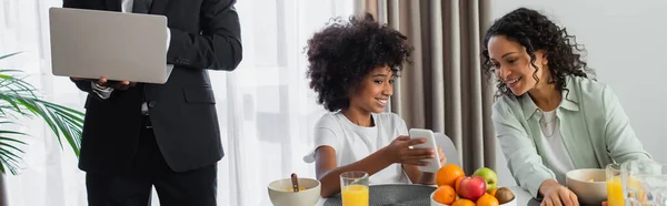 Африканский американец в костюме стоит с ноутбуком в то время как счастливая дочь показывает смартфон матери во время завтрака, баннер — стоковое фото