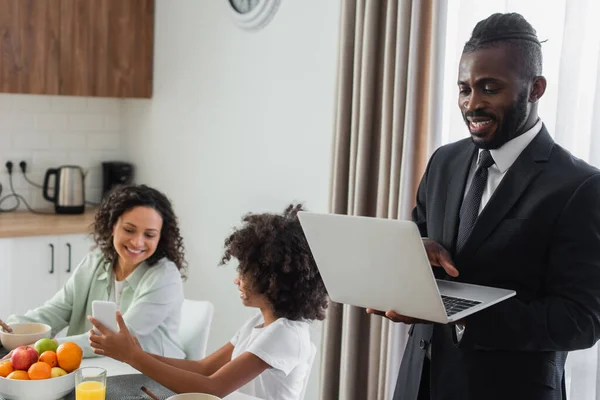 Glücklich afrikanisch-amerikanischer Mann im Anzug mit Laptop, während Tochter Mutter beim Frühstück Smartphone zeigt — Stockfoto