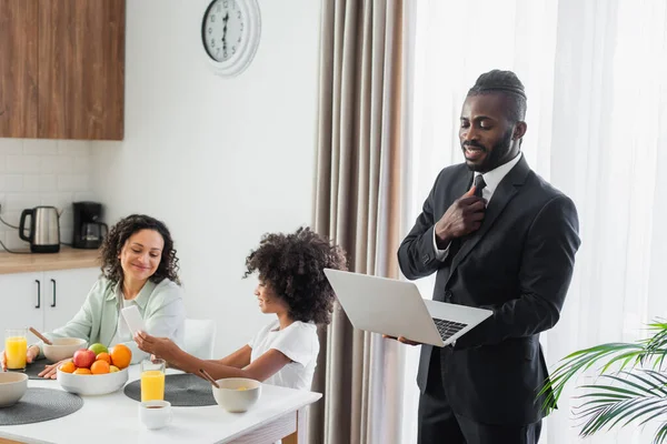 Happy african american man in suit using laptop while girl showing smartphone to mother during breakfast — Stock Photo