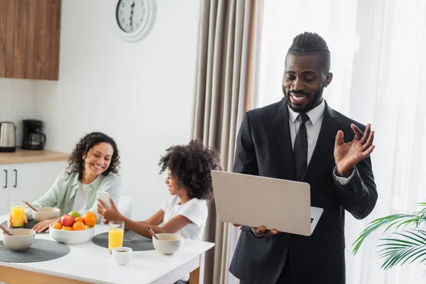 Cheerful african american man in suit using laptop while girl showing smartphone to mother during breakfast — Stock Photo