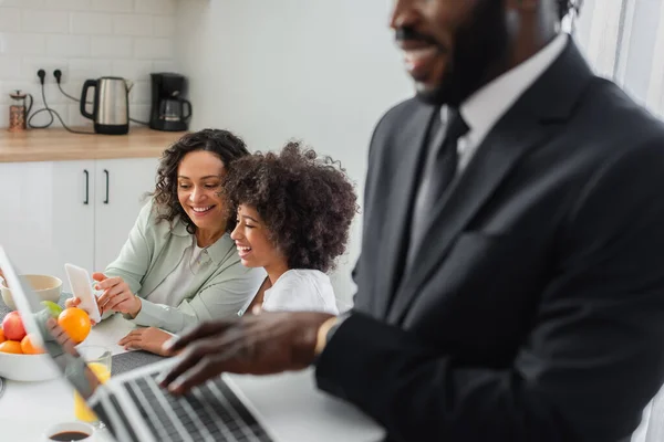 Cheerful african american mother pointing at smartphone near daughter and husband in suit using laptop on blurred foreground — Stock Photo