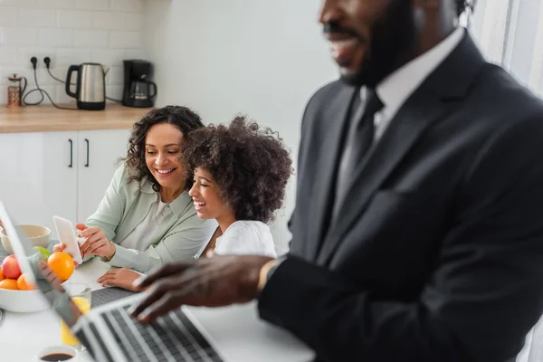 Allegra madre afroamericana che punta lo smartphone vicino a figlia e marito in giacca e cravatta utilizzando il computer portatile in primo piano sfocato — Foto stock