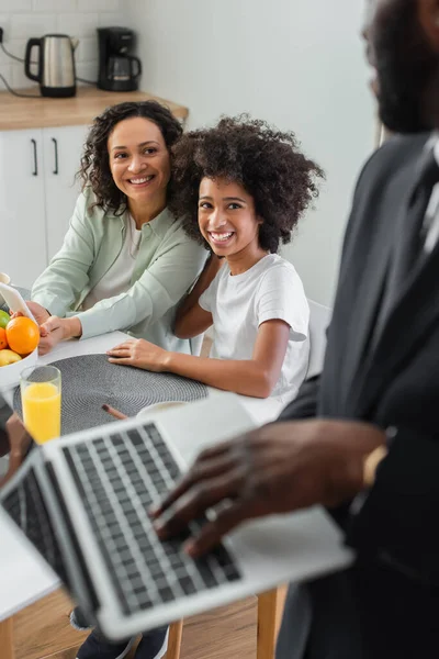 Mãe americana africana alegre segurando smartphone perto da filha e olhando para o marido de terno usando laptop em primeiro plano borrado — Fotografia de Stock