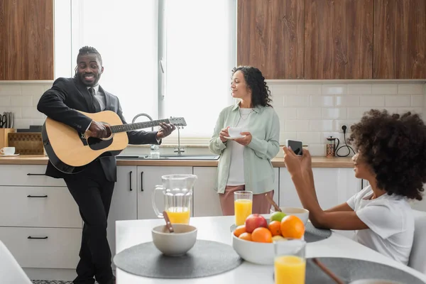 Cheerful african american girl taking photo of father in suit playing acoustic guitar near wife in kitchen — Stock Photo