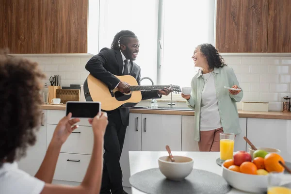 Lockiges afrikanisch-amerikanisches Kind fotografiert Vater im Anzug, der in der Küche neben glücklicher Frau Akustikgitarre spielt — Stockfoto