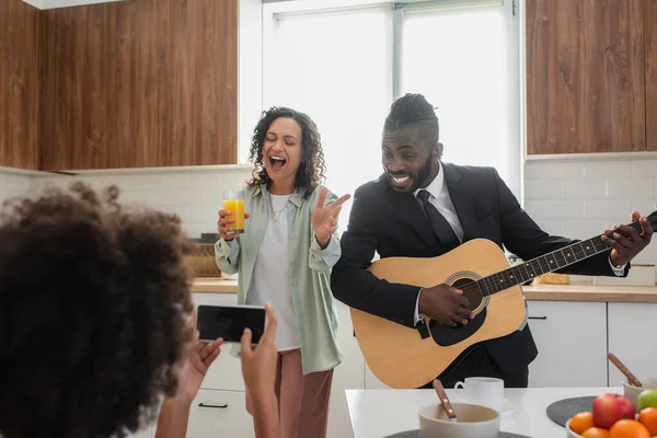 Curly african american kid taking photo of happy father in suit playing acoustic guitar near mother singing in kitchen — Stock Photo