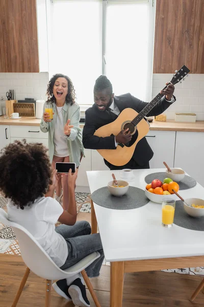 Кучерява афроамериканська дівчина фотографує щасливого батька в костюмі, граючи на акустичній гітарі біля мами співає на кухні — стокове фото