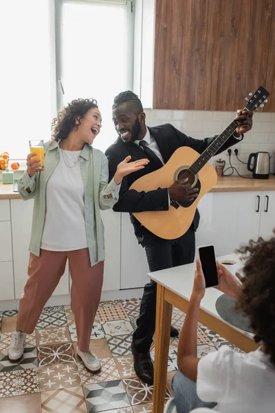 Chica afroamericana rizada tomando fotos de padres felices tocando la guitarra acústica y cantando en la cocina - foto de stock