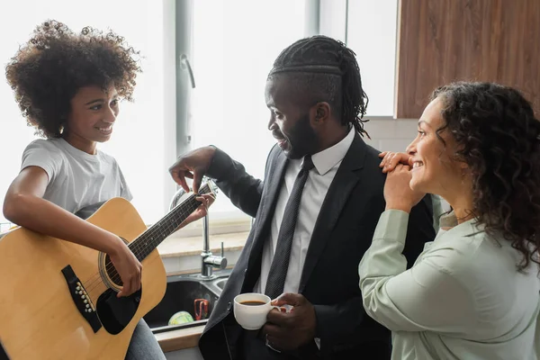 Heureux afro-américain homme avec tasse de café enseignement fille préadolescente jouer de la guitare acoustique près femme gaie — Photo de stock