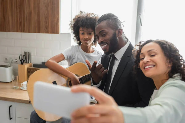 Счастливая африканская американка делает селфи с мужем и дочерью-подростком, играющими на акустической гитаре — стоковое фото
