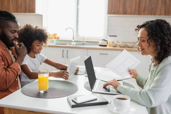 Heureuse femme afro-américaine tenant du papier près d'un ordinateur portable et la famille en utilisant une tablette numérique dans la cuisine — Photo de stock