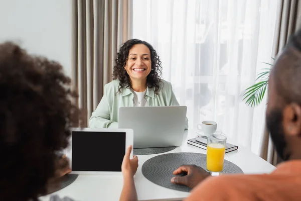 Mujer afroamericana feliz usando el ordenador portátil, mientras que el marido y el niño sentado con la tableta digital en la cocina - foto de stock