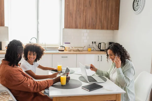 Feliz afroamericano freelancer hablando en el teléfono inteligente cerca de la computadora portátil, mientras que el marido y la hija utilizando la tableta digital en la cocina - foto de stock