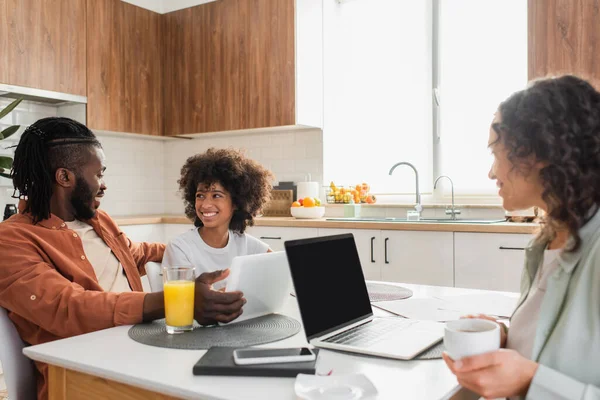 Mujer afroamericana feliz sosteniendo taza cerca de la computadora portátil mientras marido e hija hablando cerca de la tableta digital en la cocina - foto de stock