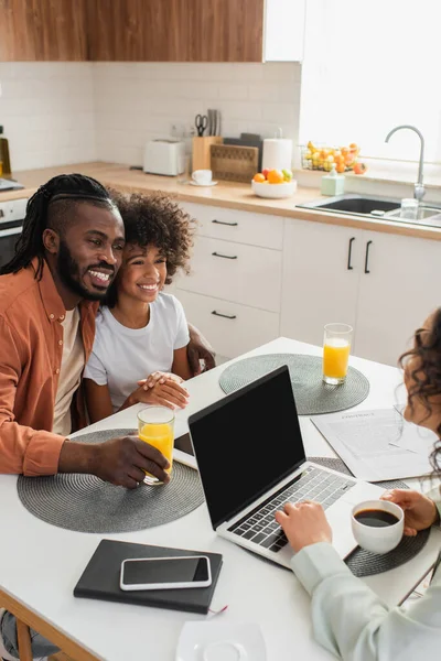 Donna afro-americana che tiene la tazza vicino al computer portatile mentre marito e figlia felici sorridono in cucina — Foto stock