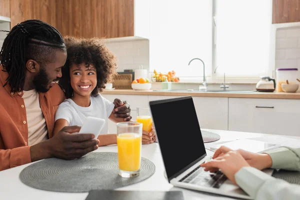 Glücklich afrikanisch-amerikanischer Vater hält Smartphone in der Hand und umarmt Tochter, während Ehefrau von zu Hause aus am Laptop arbeitet — Stockfoto