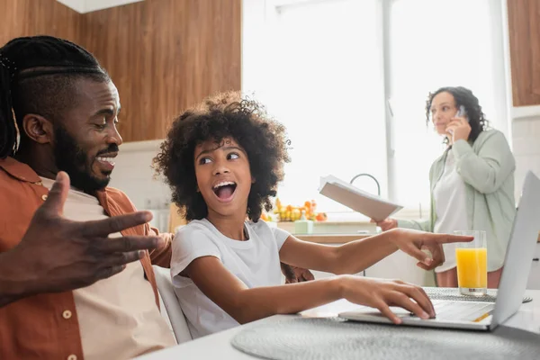 Feliz afroamericano padre mirando emocionado hija preadolescente apuntando a la computadora portátil cerca de la esposa en la cocina - foto de stock