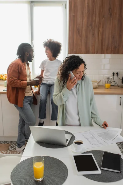 African american woman talking on smartphone and looking at documents near husband and daughter on blurred foreground — Stock Photo