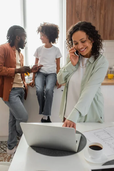 Mujer afroamericana feliz hablando en teléfono inteligente y el uso de la computadora portátil cerca de la familia en la cocina - foto de stock