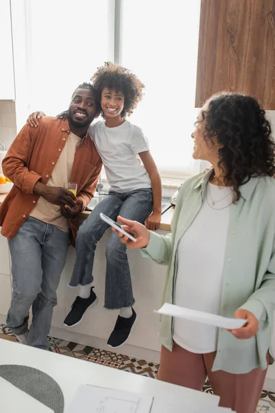 Mujer afroamericana rizada sosteniendo teléfono inteligente y papel y mirando a la familia feliz en la cocina - foto de stock