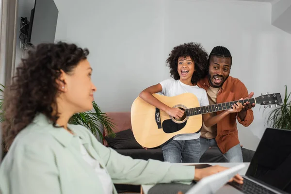 Chica afroamericana rizada tocando la guitarra acústica cerca de padre y madre trabajando desde casa - foto de stock