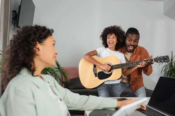 Encaracolado menina americana africana tocando guitarra acústica perto de pai e mãe trabalhando em casa — Fotografia de Stock