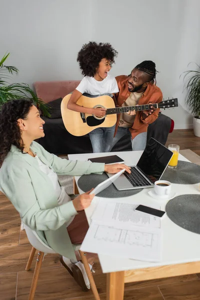 Feliz mulher afro-americana segurando tablet digital enquanto olha para a filha tocando guitarra acústica perto do marido — Fotografia de Stock