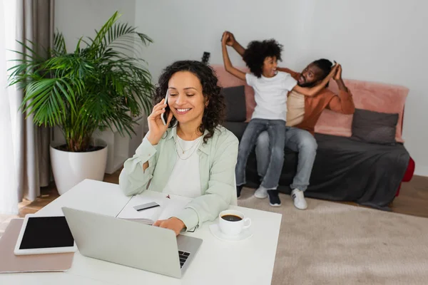 Glücklich afrikanisch-amerikanische Frau spricht auf Smartphone und benutzt Laptop nahe Familie auf verschwommenem Hintergrund — Stockfoto