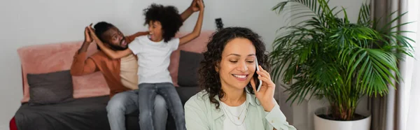 Щаслива афроамериканська жінка розмовляє на смартфоні поблизу сім'ї на розмитому фоні, банер — стокове фото