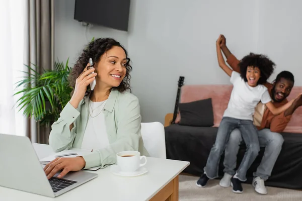 Весела афро-американська жінка говорить на смартфоні і використовує ноутбук біля щасливої сім'ї на розмитому фоні — стокове фото
