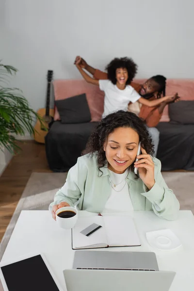 Mulher americana africana alegre falando no smartphone e segurando copo perto da família feliz no fundo borrado — Fotografia de Stock