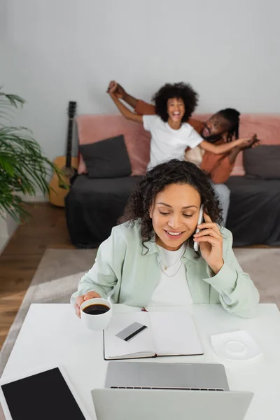 Femme afro-américaine gaie parlant sur smartphone et tenant tasse près de famille heureuse sur fond flou — Photo de stock