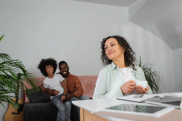 Allegra donna afro-americana in possesso di una tazza di caffè vicino alla famiglia su sfondo sfocato — Foto stock