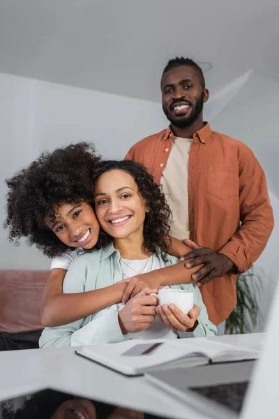 Allegra ragazza afro-americana che abbraccia la madre con una tazza di caffè vicino al padre sorridente — Foto stock