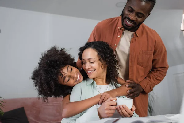 Allegra afro americano ragazza abbracciare madre con tazza di caffè vicino gioioso padre — Foto stock