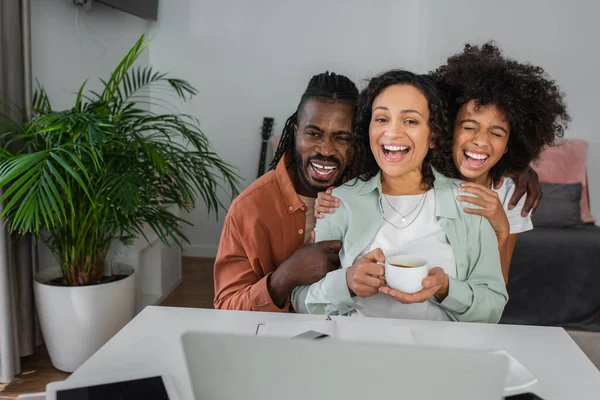 Homem americano africano alegre e mulher abraçando menina pré-adolescente com copo perto de dispositivos na mesa — Fotografia de Stock