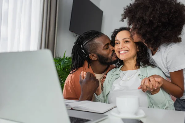 Africano americano uomo e preteen ragazza baci felice donna vicino laptop su scrivania — Foto stock