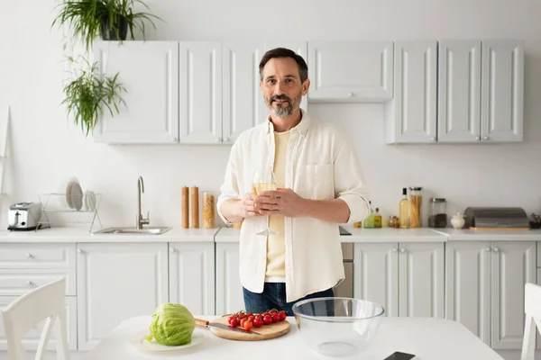 Homme souriant avec un verre de vin blanc regardant la caméra près des tomates cerises mûres et de la laitue fraîche — Photo de stock